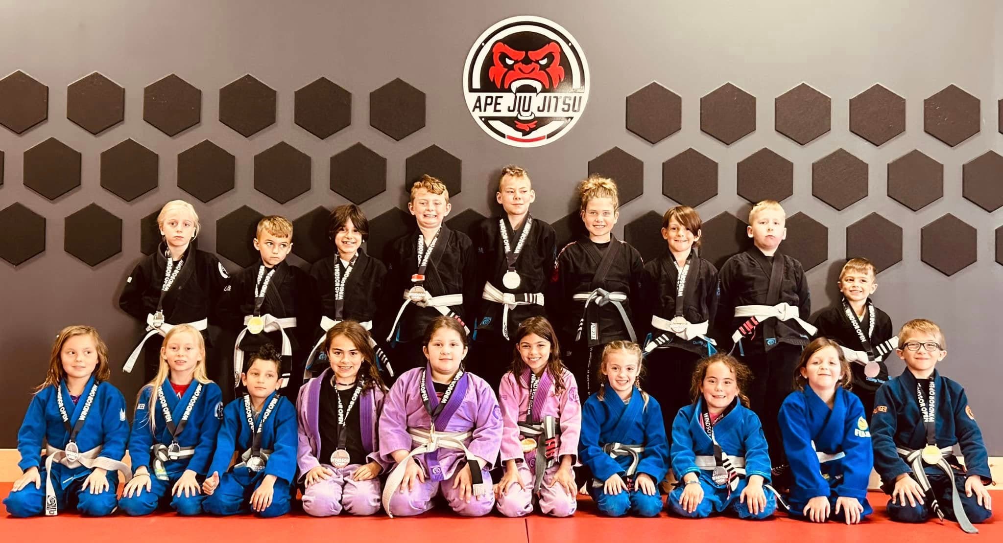 Nineteen children from Seng's in blue, black and purple gis, gathered under the Ape Jiu Jitsu crest, most of them wearing Windsor Open medals.