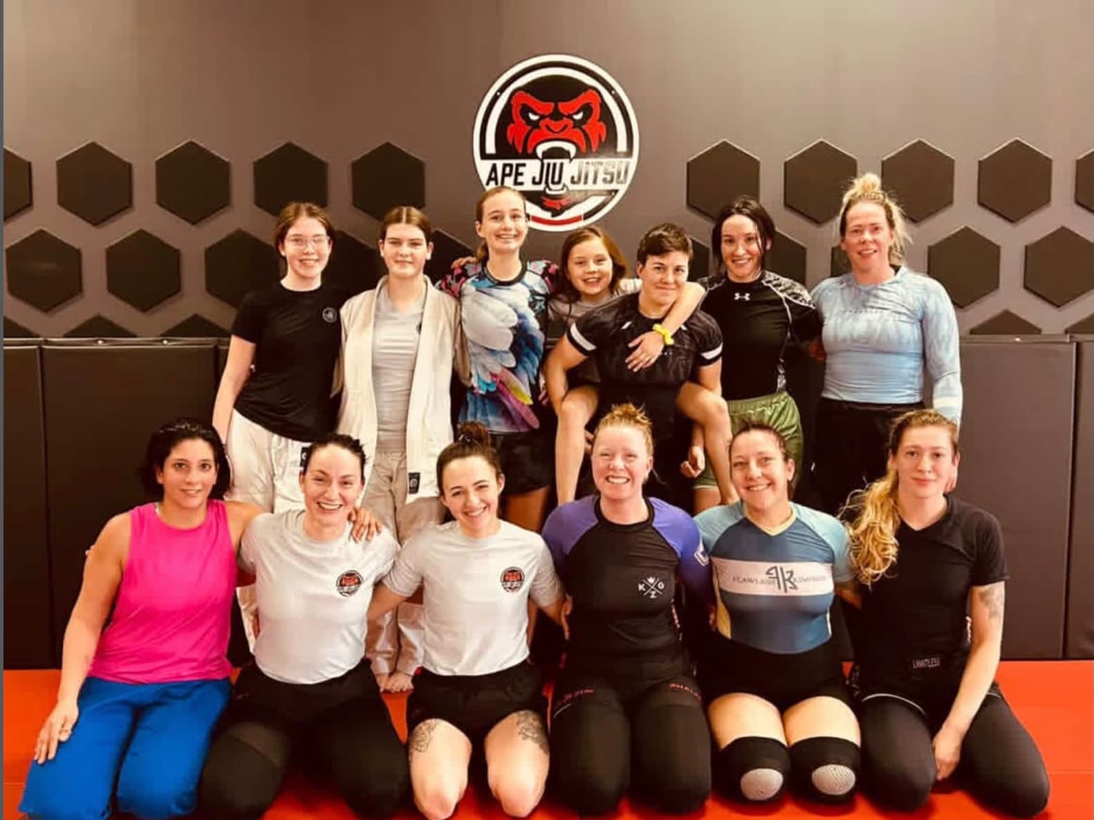 Thirteen women and girls from Seng's kneeling and standing together on the mat under the Ape Jiu Jitsu crest.
