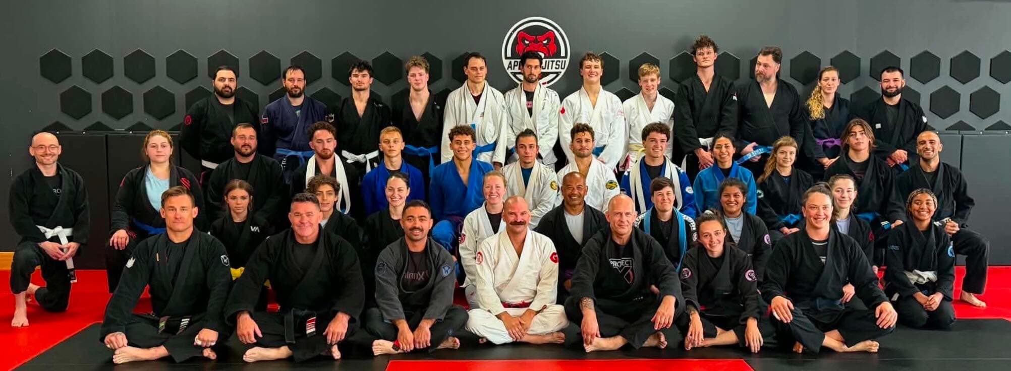 Around forty students and coaches of Seng's Martial Arts together on the mat in three rows, wearing white, blue, grey and black gis, under the Ape Jiu Jitsu crest on the wall.