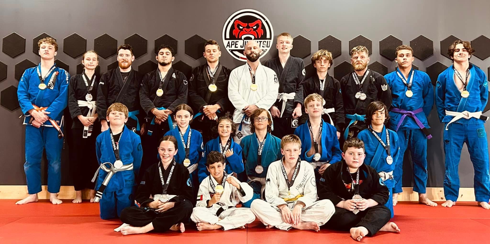 The Ape Jiu Jitsu team, children through adults, standing together under the crest in blue, black and white gis, wearing Windsor Open medals.