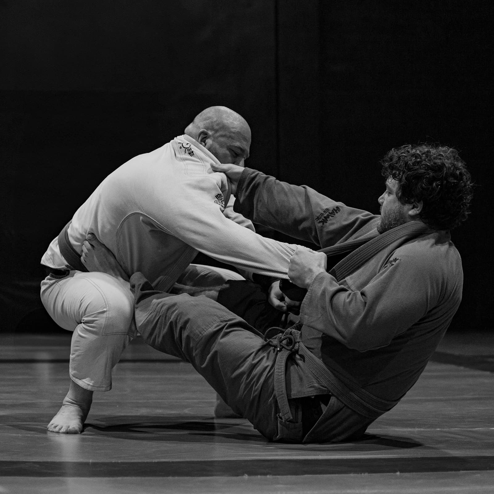 Two men in gis grappling on the mat, one working from open guard.