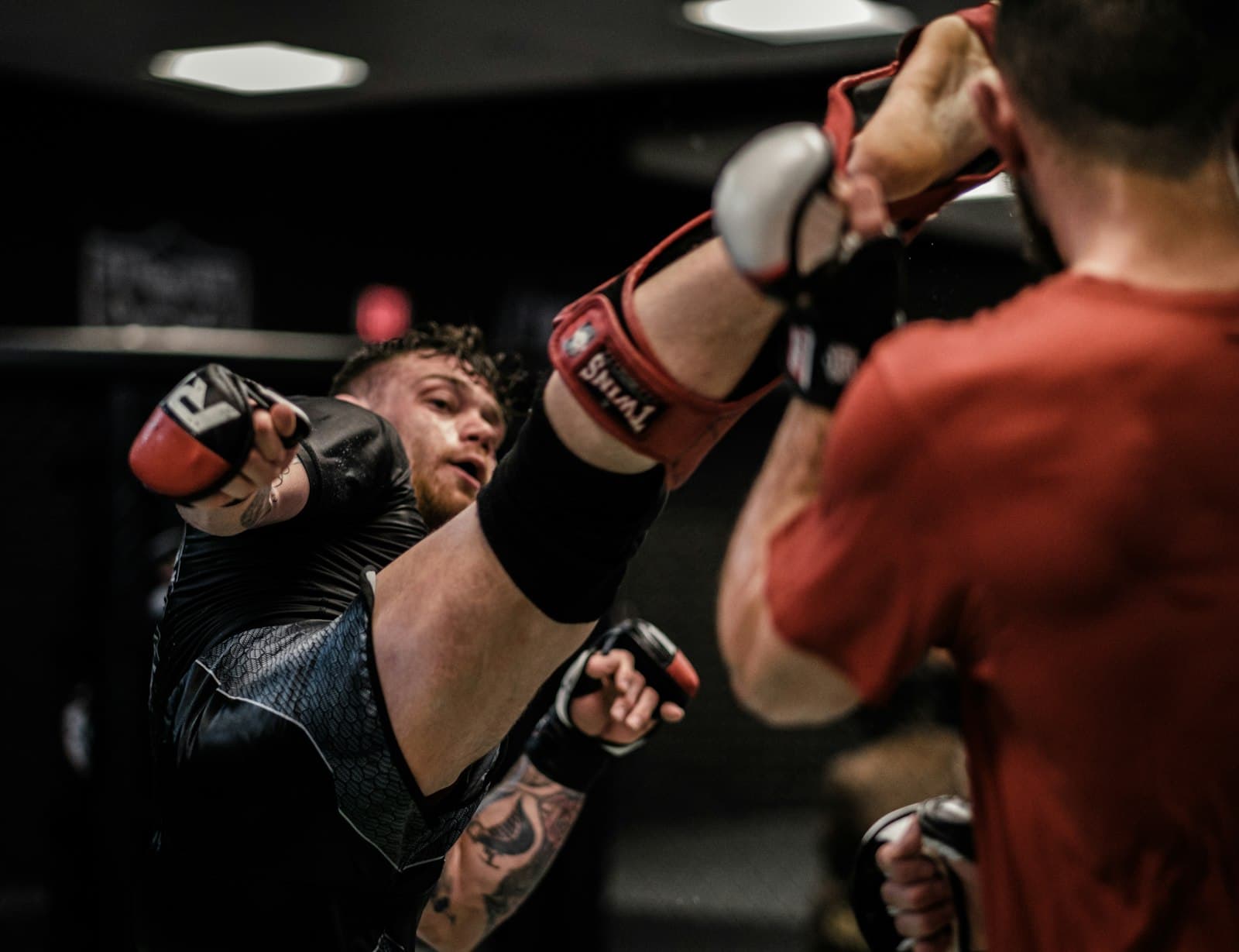 A fighter in gloves and shin pads landing a high kick on a coach holding pads.